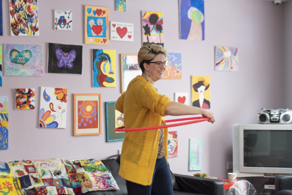 A woman with short blonde hair smiling and hula hooping with a bright pink hoop. She's wearing a bright yellow cardigan. Behind her are lots of bright paintings on a lilac wall