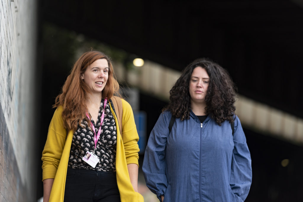 Head and shoulders shot of two people walking and talking. The person on the left is looking to the right and is wearing a yellow cardigan. The person on the right has long curly hair, is looking down and wearing a dark blue coat