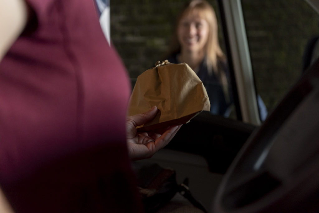 Focus is on a paper bag being passed out of a van window to someone who is standing outside , their face is blurred and out of focus