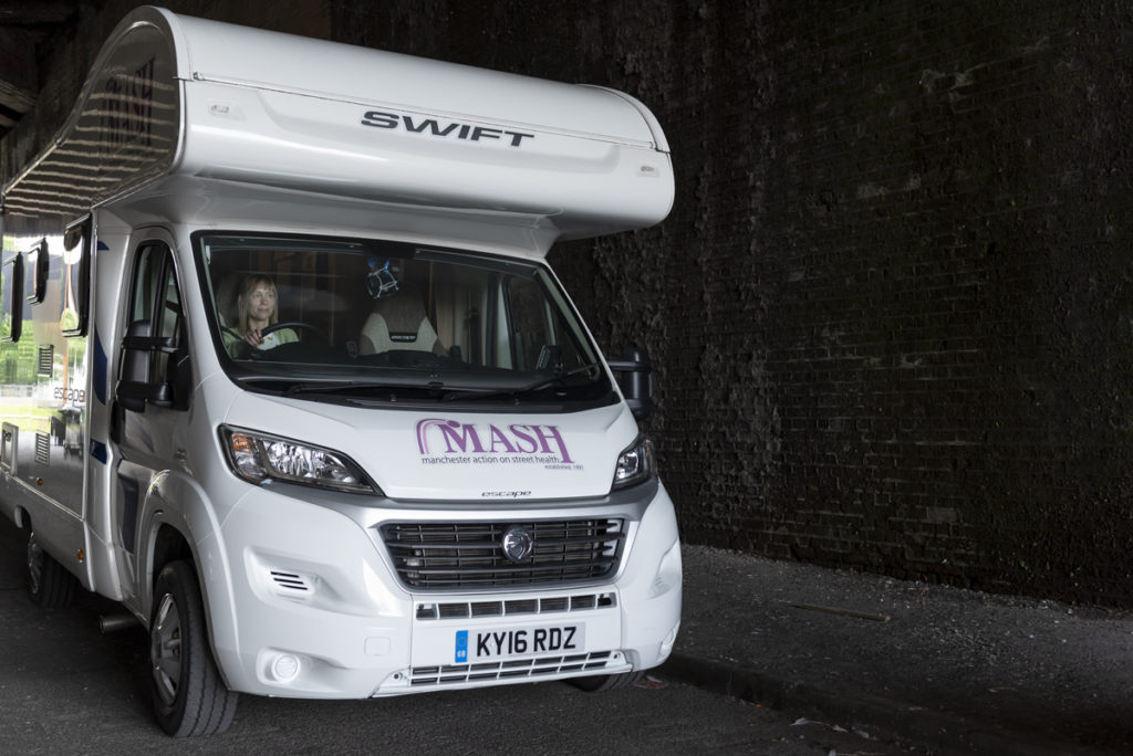 MASH van, an adapted white camper van. MASH is written across the front in purple font. It is parked up under a bridge and you can see a woman in the driver's seat