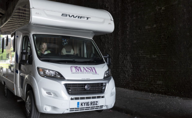 MASH van, an adapted white camper van. MASH is written across the front in purple font. It is parked up under a bridge and you can see a woman in the driver's seat