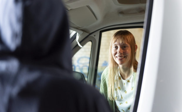 A picture of a woman with long blonde hair smiling out of a vehicle window to someone who's blurred in the left hand of the shot