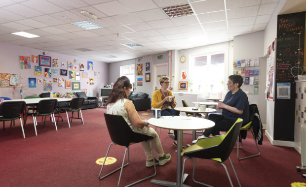 Wide shot of MASH drop-in centre. On the right three people are sat talking at a small white table. There is a lilac wall in the background displaying bright artworks by MASH service users