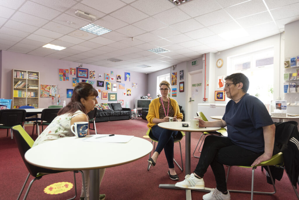 Three people sat at round tables talking. Behind them is a bright room with lots of colourful artwork on the walls. 
