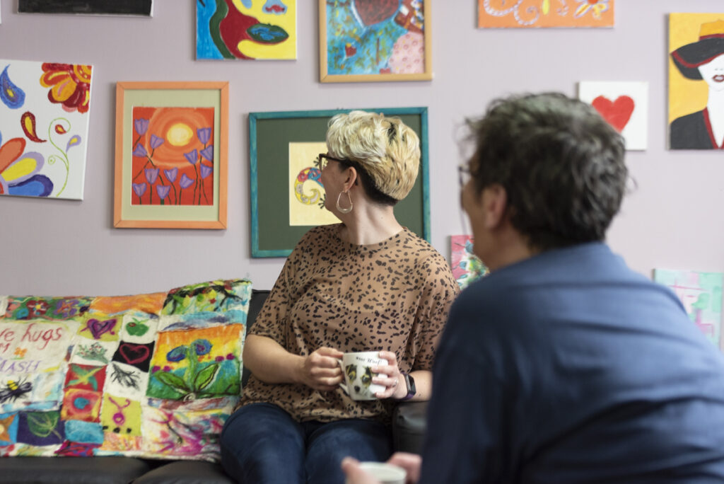 Two people have their heads turned towards bright artworks on the wall behind them. They are both holding mugs