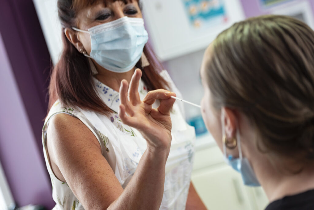 A woman wearing a blue medical mask is taking a swab sample from someone's mouth. You can just see the back of their head. Dark hair tied up
