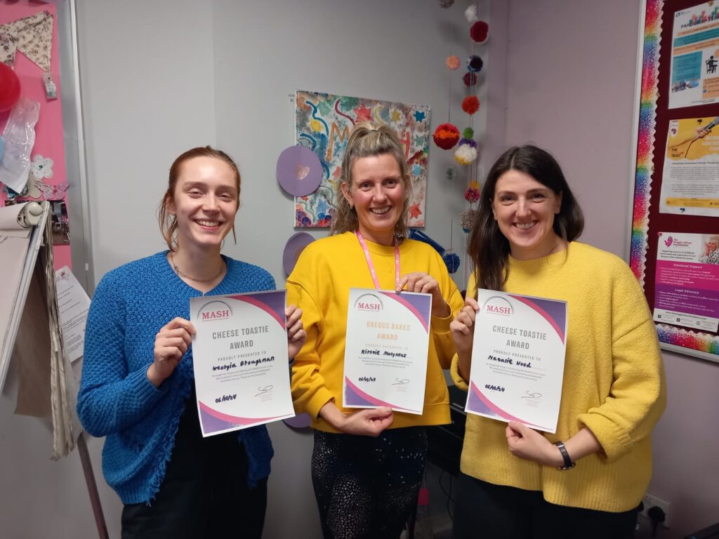 Three volunteers stood  with colourful picture on the wall behind them, holding certificates up smiling.