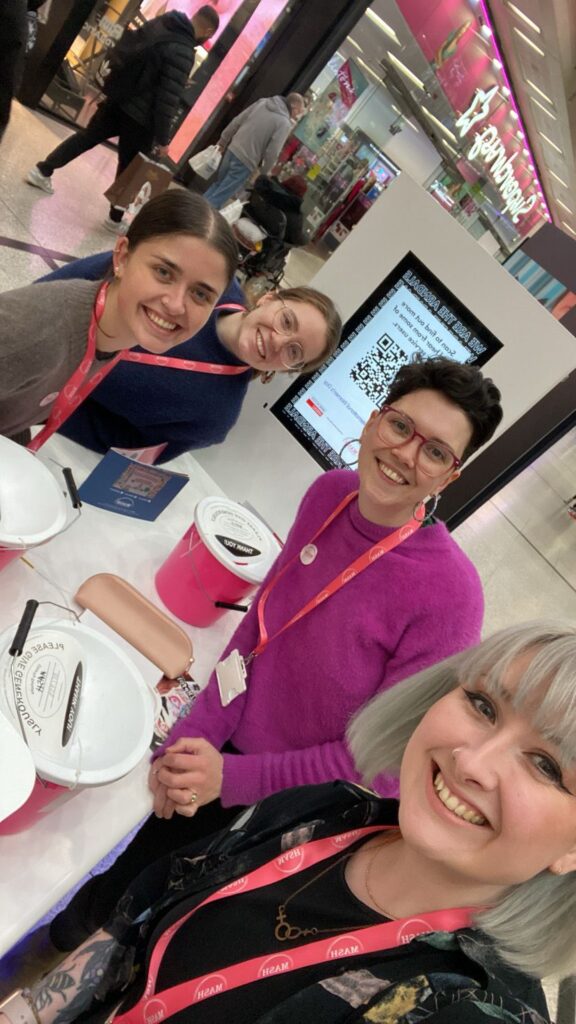 Fundraiser, Janelle, and three volunteers sat down showing heads and shoulder smiling at the camera with their pink lanyards at the Arndale