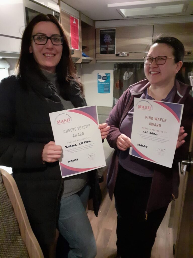 Two volunteers stood smiling at the camera holding their certificates in the MASH van.